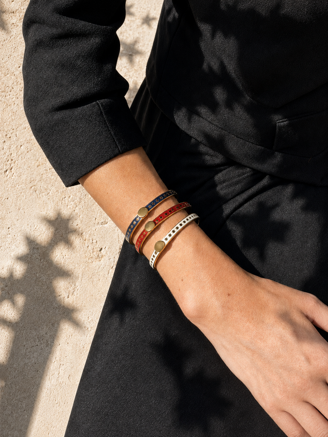 A close-up of a person's wrist wearing four stacked bracelets, including a blue and red woven bracelet with gold-toned round beads and a white beaded bracelet with gold-toned dots, against a black dress and textured beige wall with leaf shadows.